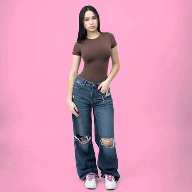 Woman in a brown shirt and ripped jeans wearing white sneakers with Pretty Pink elastic shoelaces on a pink background.